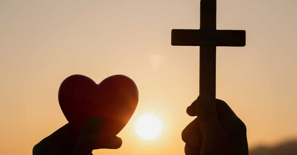 Silhouette Hand Holding Heart Shape And Religious Cross During Sunset