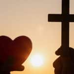 Silhouette Hand Holding Heart Shape And Religious Cross During Sunset