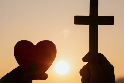 Silhouette Hand Holding Heart Shape And Religious Cross During Sunset